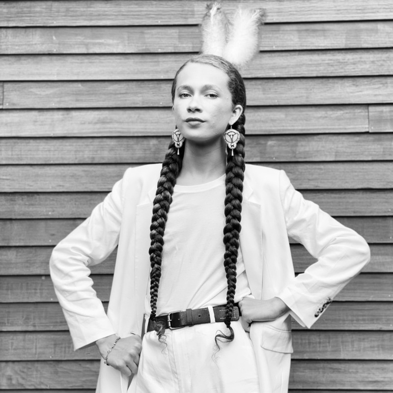 Image of Sherenté Harris with hands on hips, staring at the camera in front of wood siding