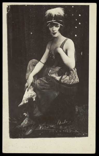 Download the full-sized image of Chris Bennett in drag sits on stage, wearing a feathered head garment. Process print, 1921.