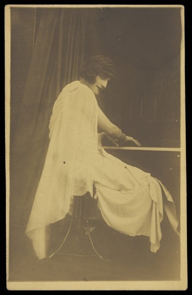 Download the full-sized image of A man wearing a white dress sits playing the piano. Photographic postcard, 192-.