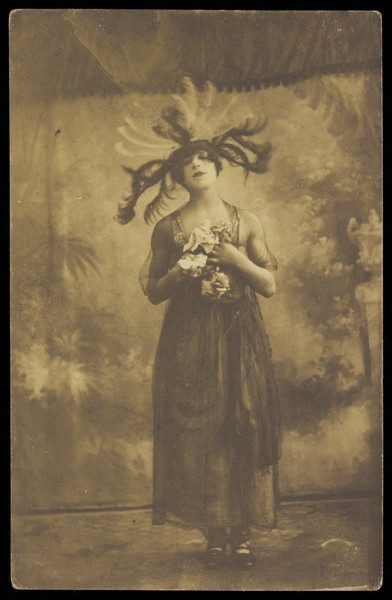 Download the full-sized image of A performer in drag poses on stage wearing a head garment with sprawling feathers. Photographic postcard, 191-.