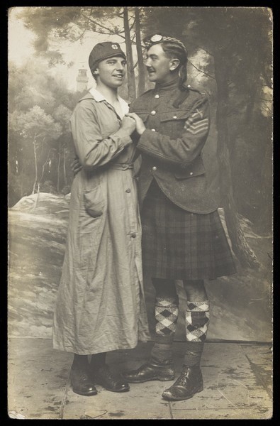 Download the full-sized image of Two soldiers playing in a military concert party embrace: one is in drag and the other is wearing a kilt. Photographic postcard, 191-.