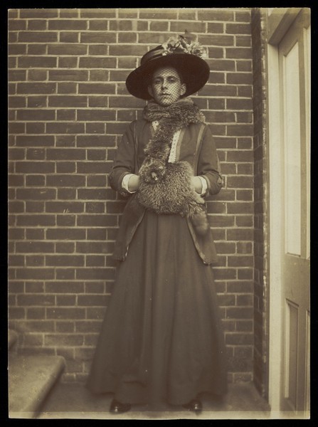 Download the full-sized image of A man in drag, posing in a dark hat in front of a brick wall. Photographic postcard, ca. 1910.
