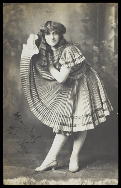 Download the full-sized image of An amateur actor in drag holds up his impressive folded dress to one side. Photographic postcard, 191-.