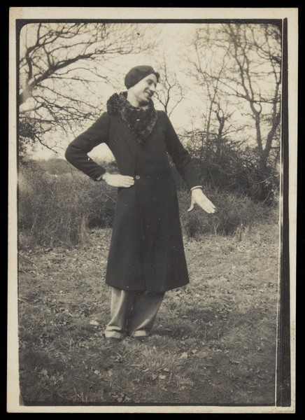 Download the full-sized image of A man in drag poses on some grass, wearing a long thick coat. Photograph, 1934.
