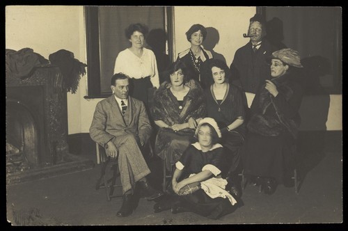 Download the full-sized image of Amateur actors, some in drag, gather for a group portrait, in a room with a fireplace. Photographic postcard, 192-.