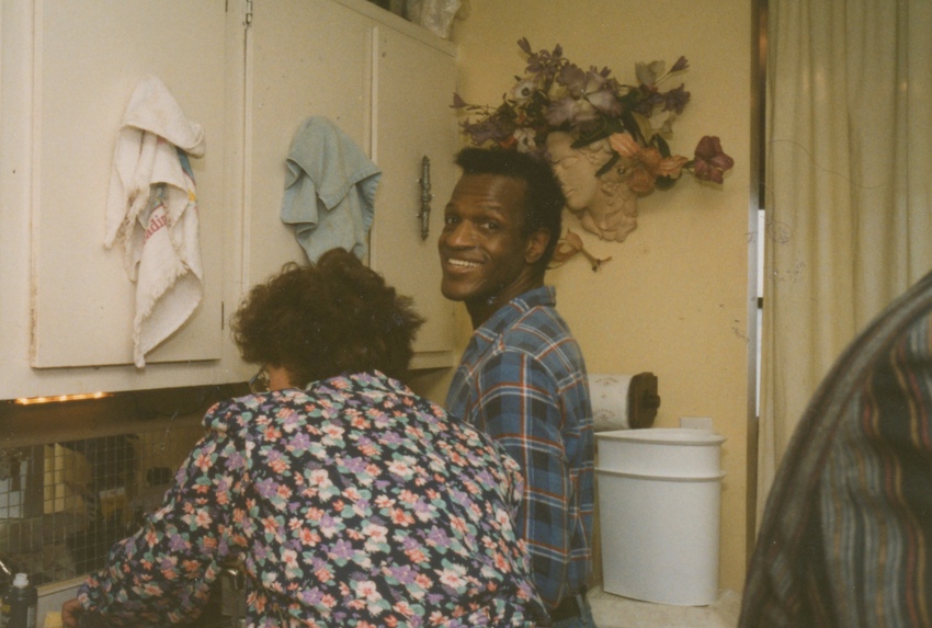 Download the full-sized image of A Photograph of Marsha P. Johnson in a Yellow Kitchen Wearing a Blue Flannel Shirt, Smiling Over Her Shoulder