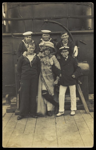 Download the full-sized image of Six sailors, one in drag, pose for a group portrait on the deck of H.M.S. St. George. Photographic postcard, 191-.