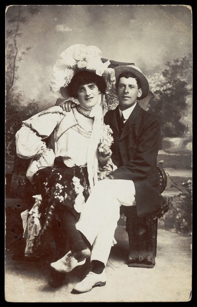 Download the full-sized image of Two men, one in drag, sit together on a bench. Photographic postcard, 190-.