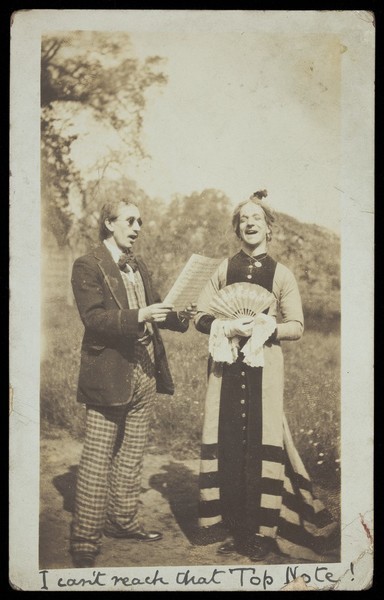 Download the full-sized image of Harry Hawksbee in drag, rehearsing a song with a man wearing sunglasses; posing outdoors. Photographic postcard, 1914.