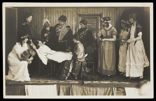 Download the full-sized image of Amateur actors, some in drag, on stage attending to the death of a female character. Photographic postcard, 191-.