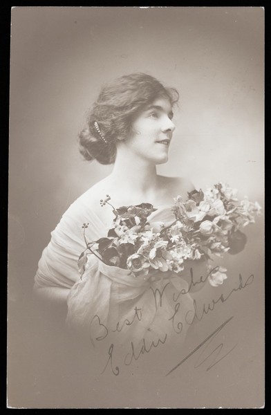 Download the full-sized image of Eddie Edwards, an actor in drag, wearing a white fabric robe, holding a bouquet of flowers. Photographic postcard by L.E. Muller, 191-.