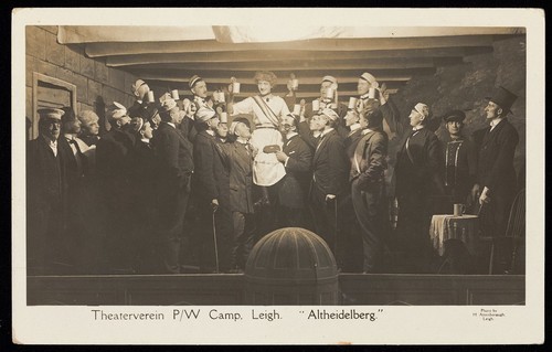 Download the full-sized image of German prisoners of war, one in drag, propose a toast during a performance at Leigh prisoner of war camp, Lancashire. Photographic postcard by H. Attenborough, 191-.