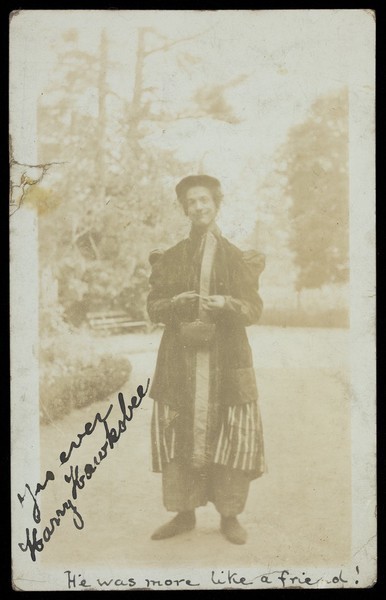 Download the full-sized image of Harry Hawksbee in drag, wearing a large coat; posing outside near trees. Photographic postcard, 1914.