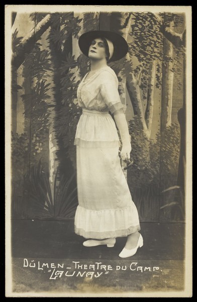 Download the full-sized image of A prisoner of war acting in an internment camp in Dülmen, performing in drag, wearing a white dress and a dark hat. Photographic postcard, 191-.