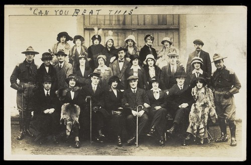 Download the full-sized image of Actors, some in drag, pose for a group portrait, in front of a large wooden doorway. Photographic postcard, 191-.