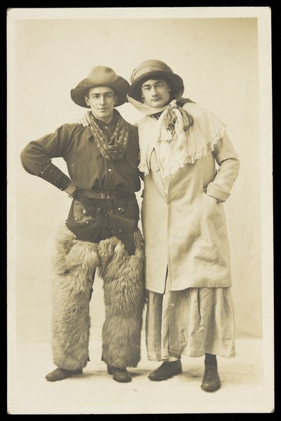 Download the full-sized image of Two men, one in drag, the other dressed as a cowboy, pose in front of a plain background. Photographic postcard, 191-.