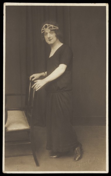Download the full-sized image of A performer in drag, wearing a black dress and tiara, leaning on a chair. Photographic postcard by W. Aspden, 1924.