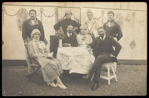 Download the full-sized image of German soldiers, some in drag, pose round a table at a concert party. Photographic postcard, 191-.