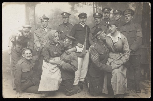 Download the full-sized image of British soldiers, two in drag, pose for a group portrait, in front of a detailed backdrop. Photographic postcard, 191-.