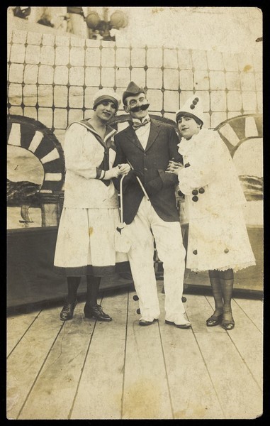Download the full-sized image of Three sailors, two in drag wearing pierrette costumes, pose on deck. Photographic postcard, 191-.