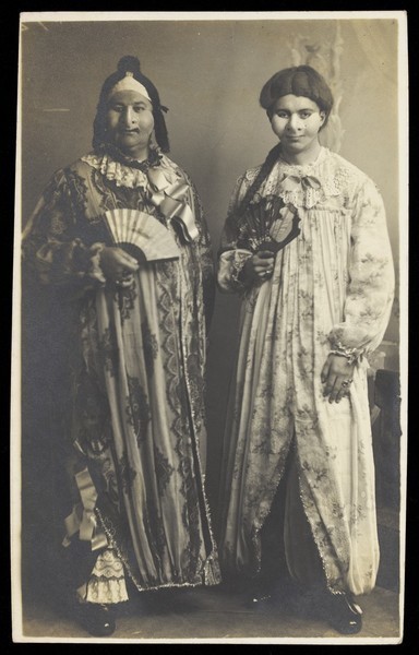Download the full-sized image of Two men in drag wearing face masks and patterned robes, holding fans. Photographic postcard, ca. 1910.