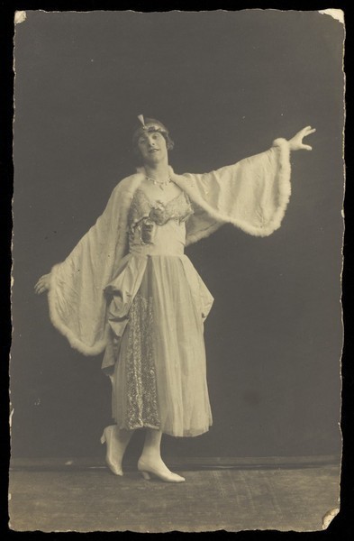 Download the full-sized image of A prisoner of war in drag, wearing a dress and white cloak, poses mid-movement with arms outstretched. Photographic postcard by Albert Melzer, 191-.