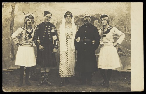 Download the full-sized image of British prisoners of war, some in drag, posing on stage arm in arm at a prisoner of war camp in Cottbus. Photographic postcard by P. Tharan, 191-.