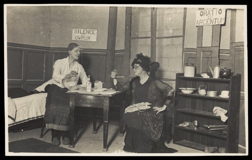 Download the full-sized image of Two amateur actors, both in drag, having a drink on stage. Photographic postcard, 191-.