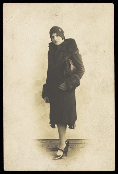 Download the full-sized image of A man in drag (?) poses wearing a long coat with fur trim and a hat with a brooch. Photographic postcard, 192-.