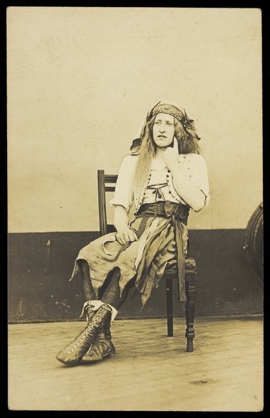Download the full-sized image of A man in drag sits pensively on a chair, on the deck of a ship. Photographic postcard, 191-.