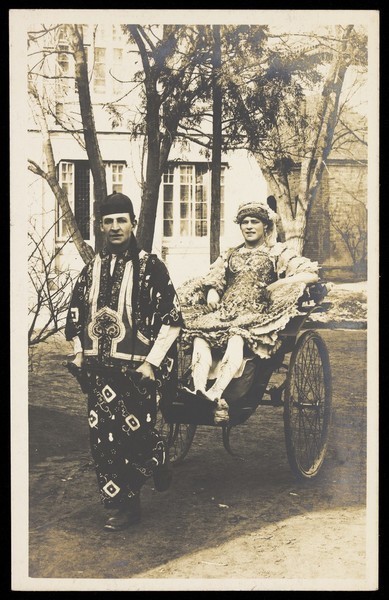 Download the full-sized image of A man in ornate drag sits in a dog-cart and is pulled along the road by a man dressed in Chinese patterned robes and hat. Photographic postcard, 191-.