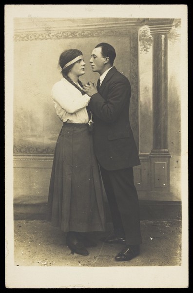 Download the full-sized image of A man in drag and a man in male clothes looking into each others' eyes. Photographic postcard.