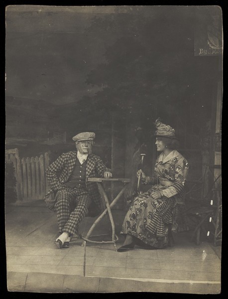 Download the full-sized image of Two sailors, one in drag, sit on stage at a table, in front of very detailed scenery. Photographic postcard, 191-.