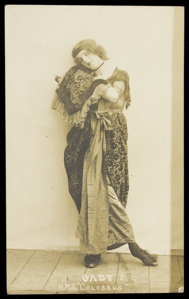 Download the full-sized image of An sailor poses in drag, wearing heavy fabric and a headband. Photographic postcard, ca. 1915-1916.