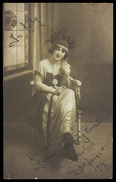Download the full-sized image of A man in drag (Charlie) sits face-on, wearing detailed costume, with a window painted on the backdrop. Photographic postcard, 1919.