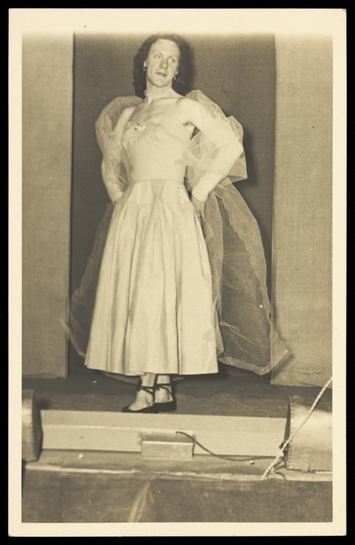 Download the full-sized image of A man in drag standing on a stage. Photographic postcard, 195-.