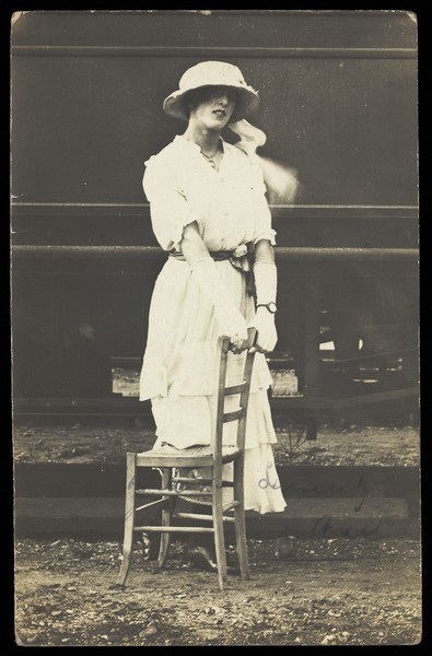 Download the full-sized image of An soldier in a concert party poses in drag, wearing white and holding a chair, standing in front of a train. Photographic postcard, 191-.