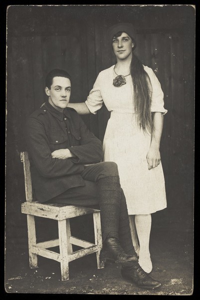 Download the full-sized image of Two prisoners of war, one in drag, pose with a chair. Photographic postcard, 191-.