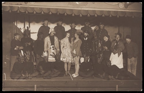 Download the full-sized image of Soldiers, some in drag, pose on stage; two men in drag face each other in the centre. Photographic postcard, 191-.