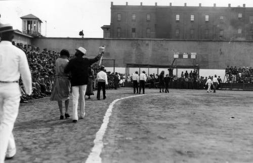 Download the full-sized image of Parade of prisoners, some in female dress, with prison buildings in background, San Quentin Little Olympics Field Meet, 1930