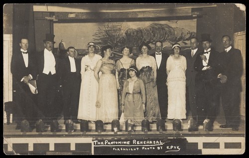 Download the full-sized image of Amateur actors, some in drag, posing on stage for a group portrait. Photographic postcard by E. Pye, 191-.