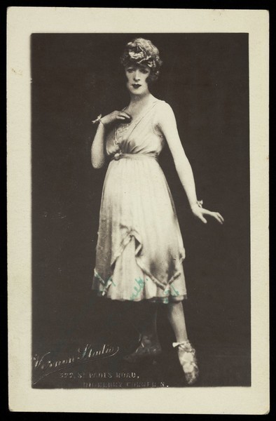 Download the full-sized image of An actor in drag wearing a light dress and styled hair, poses in front of a black background. Photographic postcard by Vernon Studio, 1921.