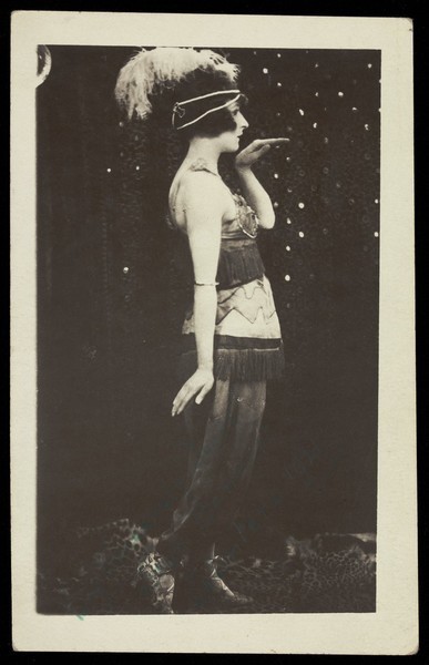 Download the full-sized image of An actor in drag, poses mid-dance, wearing a feathered head garment and a tassled dress. Photographic postcard, 1921.