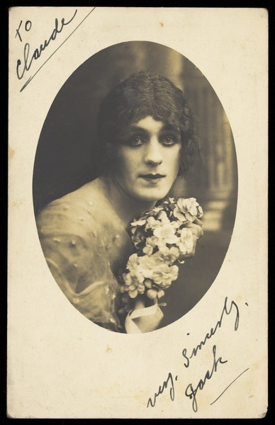 Download the full-sized image of A man in drag wears make-up and is holding flowers, posing for his portrait. Photographic postcard, ca. 1918.