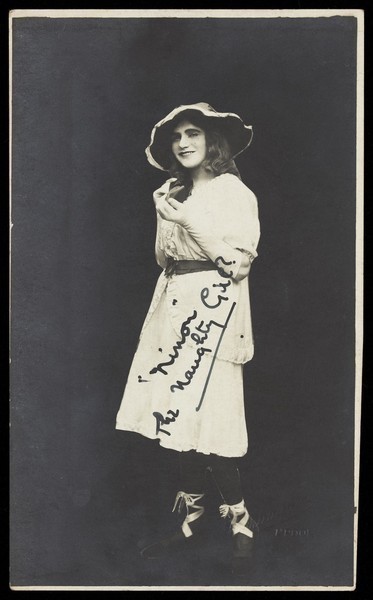Download the full-sized image of An amateur actor in drag, posing as as 'Ninon', wearing a white dress and hat. Photographic postcard, 192-.