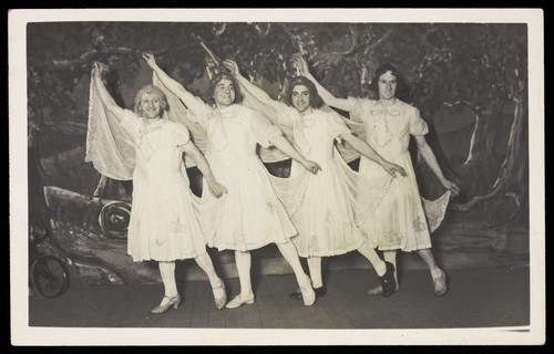 Download the full-sized image of Amateur actors in drag, wearing white dresses and dancing on stage. Photographic postcard, 191-.