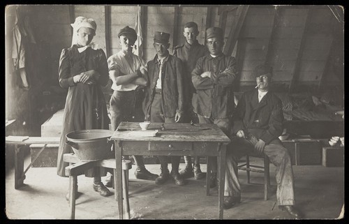 Download the full-sized image of French prisoners of war, one in drag, gather round a small wooden table. Photographic postcard, 191-.