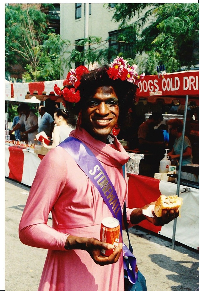 Download the full-sized image of A Photograph of Marsha P. Johnson Holding a Sandwich and Soda, Wearing a Pink Dress and Purple “Stonewall” Sash