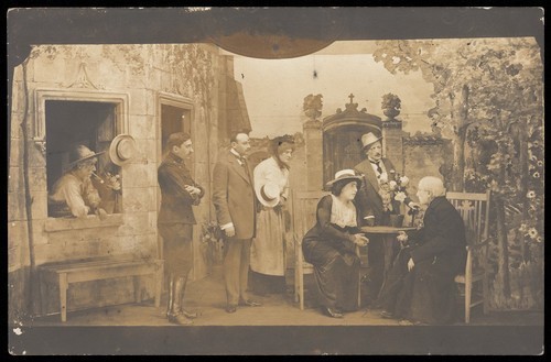 Download the full-sized image of Actors, some in drag; with the attention focused on a character sitting at a table. Photographic postcard, 191-.